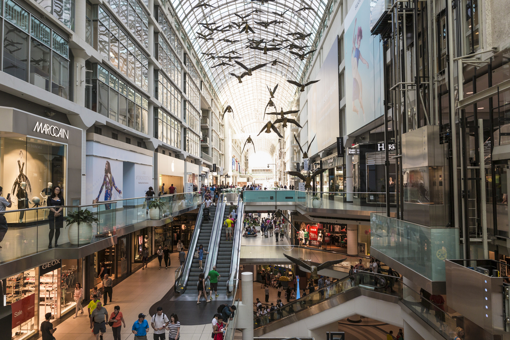 A busy modern architecture glass mall with bird sculptures hanging by the arch ceiling at the Entertainment and Fashion District, one of our picks on the best places to stay in Toronto.