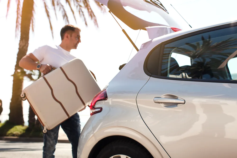 Man loads a white suitcase into a white car as he prepares for a domestic travel trip within the US as a palm tree is seen in the background