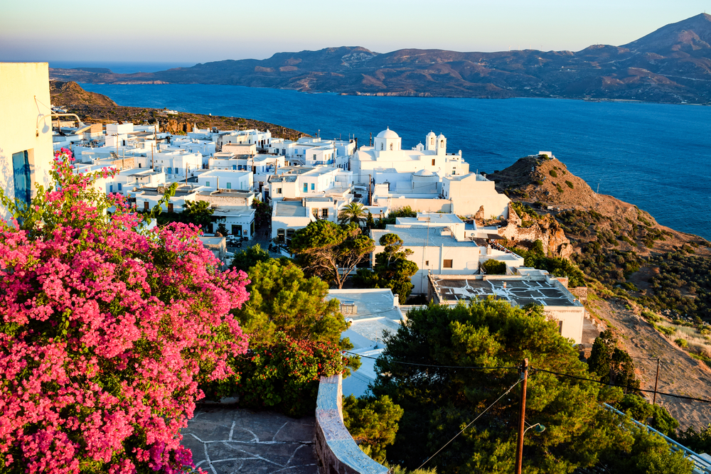 A view of a coastal village from a balcony with blooming flowers, where structure are seen to be all-white and the opposite island have tall mountains, snapped as a section image for a travel guide about safety in visiting Milos.