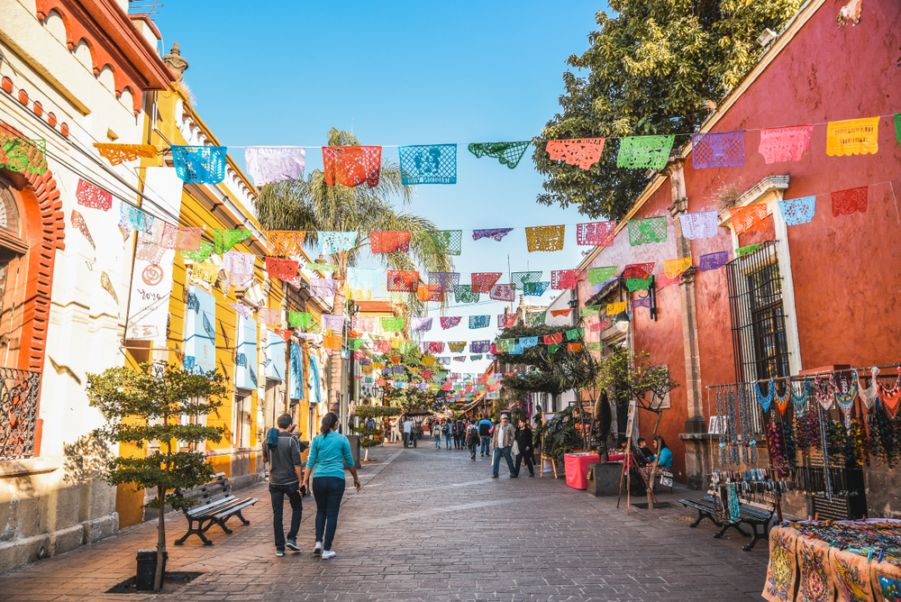 Photo of people walking through the streets of Tlaqepaque for a guide with its stone streets and colorful pink and yellow buildings and party flags above the street pictured for a guide titled Is Guadalajara Safe to Visit