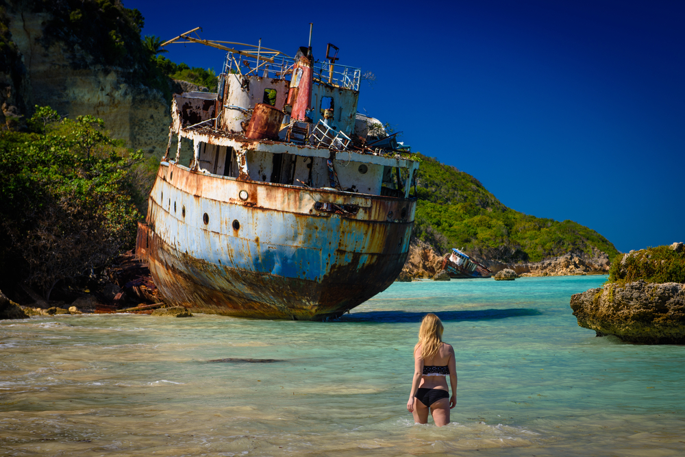 A women in her swimwear walking towards a shipwrecked boat on a rocky shore. 