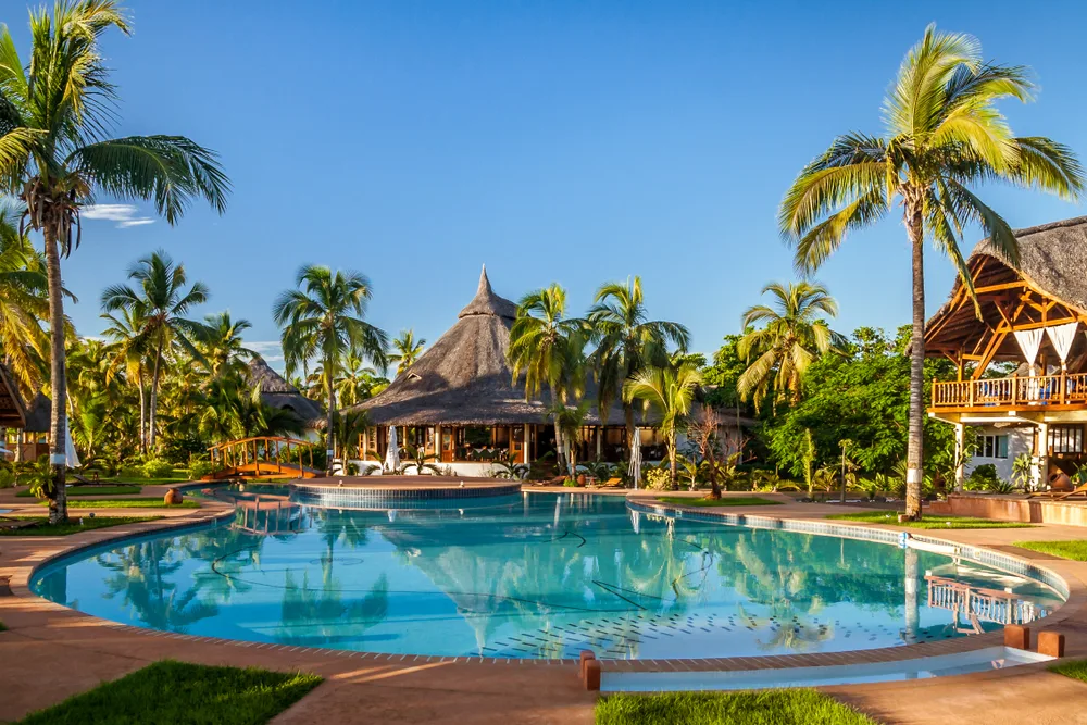 A tropical resort integrated with native structures and palm trees, a large swimming pool as at the center of the property, an image for an article about trip cost to Madagascar. 