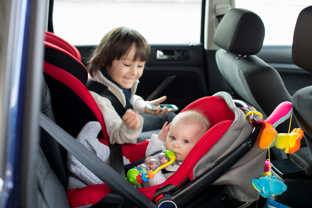 An infant and a toddler placed on a car seat while preparing for a trip, an image for a travel guide on how to travel with a car seat.