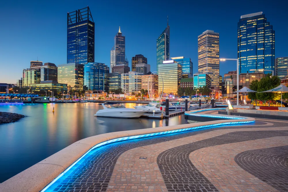 A small pier with boats and a paved walking area, in background is a city skyline at dusk, an image for a travel guide about trip cost to Australia.