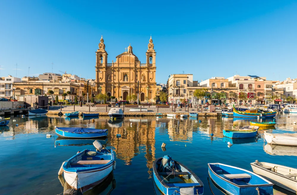 View from the lake with small fishing boats Sliema, our pick on the best areas to stay in Malta, and an old church and several city structure. 