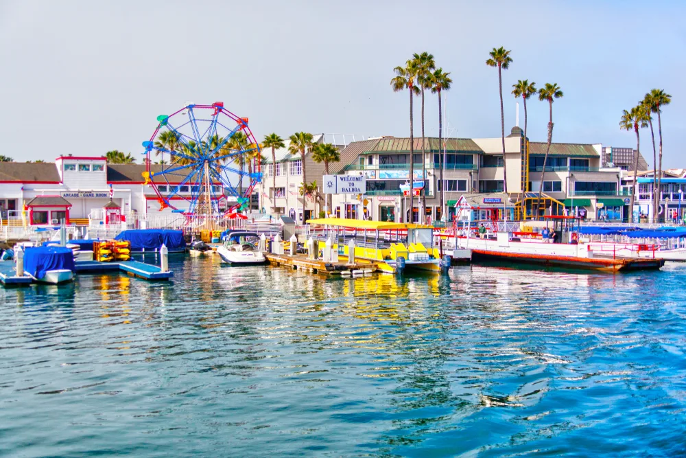 Several boats can be seen on a port with a small ferris wheel and a few tall palm trees, and some commercial structures. 