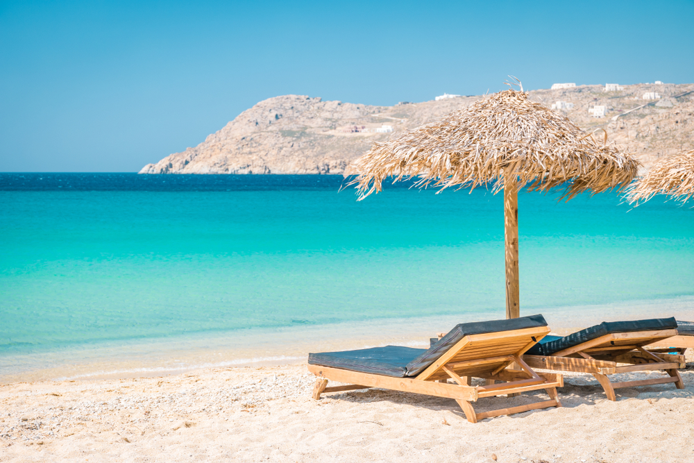 Two sunbeds is placed under a native-style beach umbrella on a beach with calm emerald waters, a section image for an article about trip cost to Mykonos.