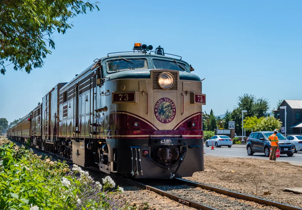 A train standby on its tracks on a countryside, and some automobiles are parked near the train, an image for an article about trip cost to Napa Valley.