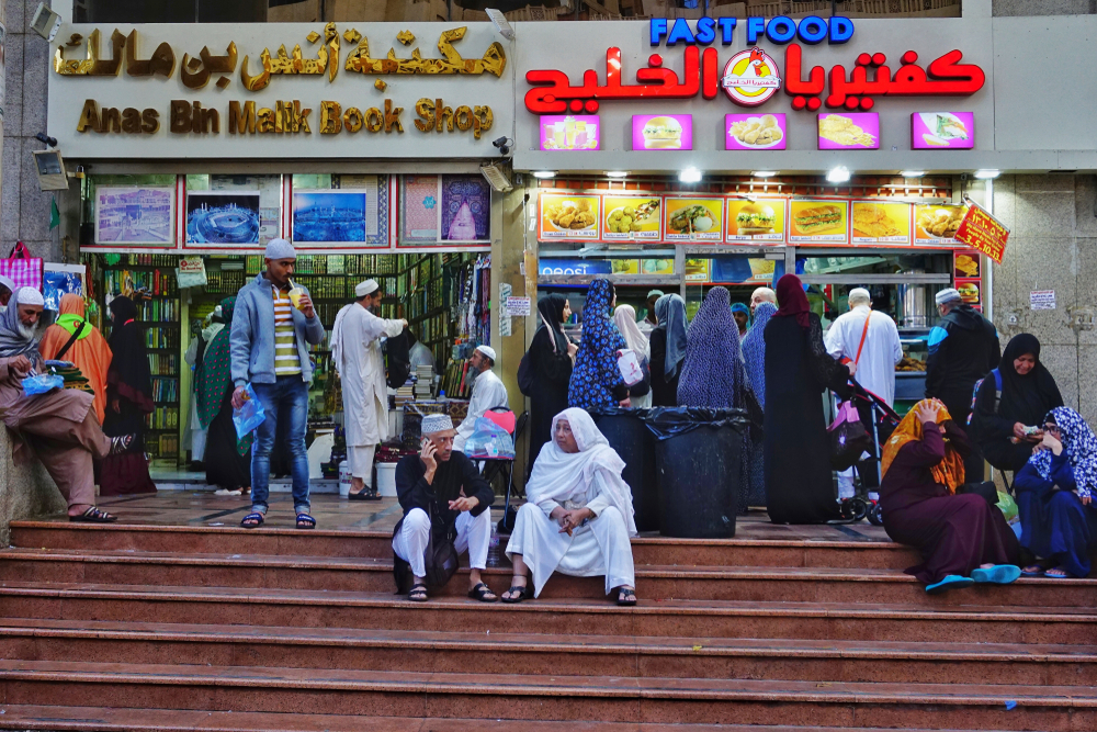 People waiting while placing an order in an express food counter beside the street, others are sitting on the stairs, an image for an article about trip cost to Mecca.