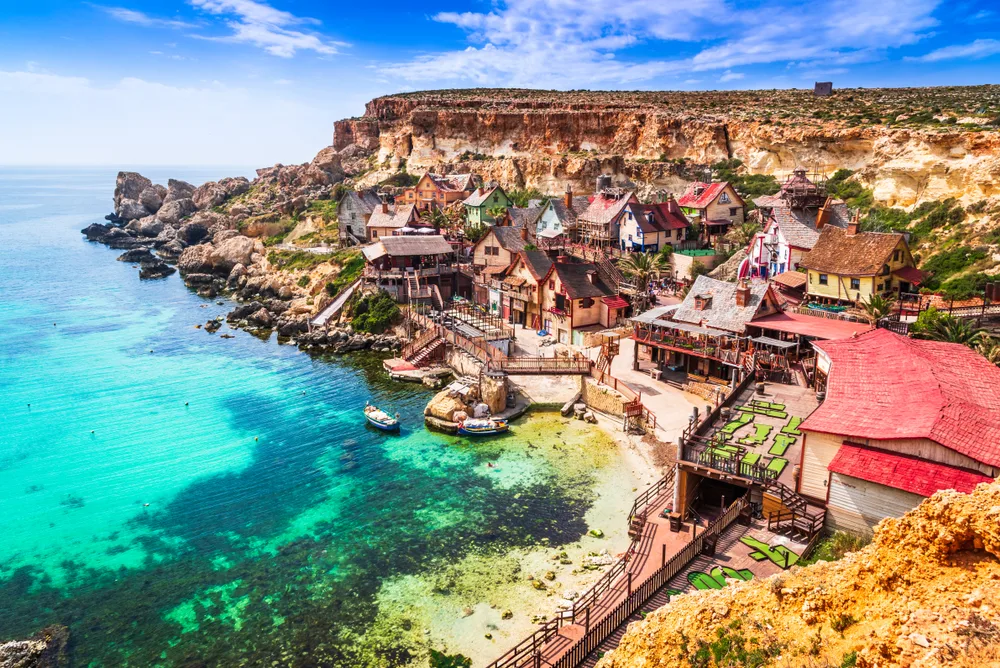 a town on a rocky coastal area in Mellieha Bay, our pick on the best areas to stay in Malta, old structures stand near the rocks and boats are docked on a pier with clear waters.