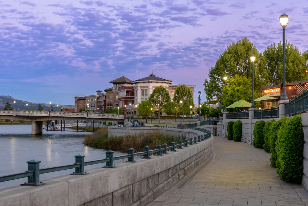 A winding river walk towards a bridge spanning the wide rider, and a large building, snapped during a cloudy late afternoon for a section image on an article about trip cost to Napa Valley.
