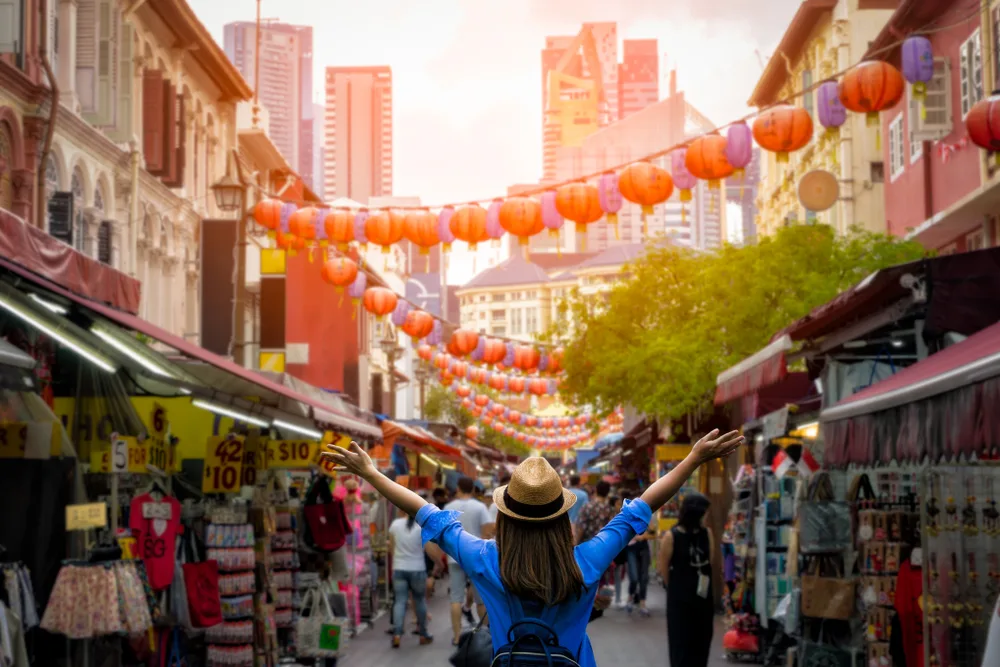 A woman wearing blur shirt and a hat spreading her arms as she enter Chinatown, and tall buildings can be seen in background. 