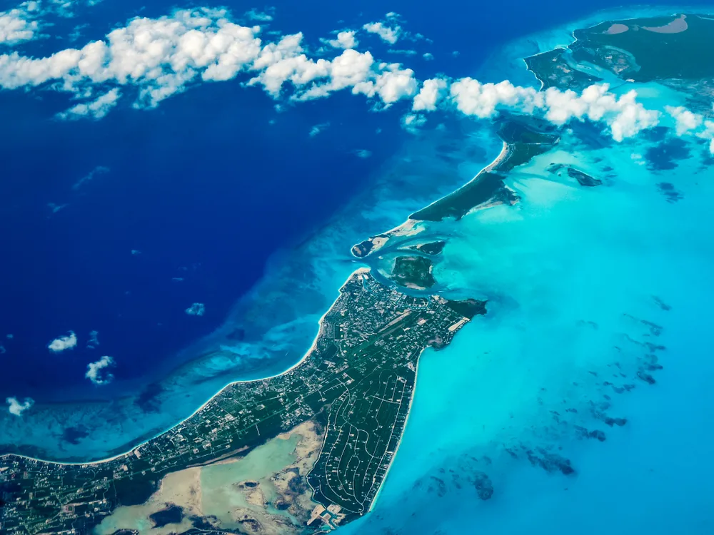 Aerial view from an airplane over Turks and Caicos Islands with a string of white clouds below the plane for a section explaining how long is a flight to Turks and Caicos with 1 stop?