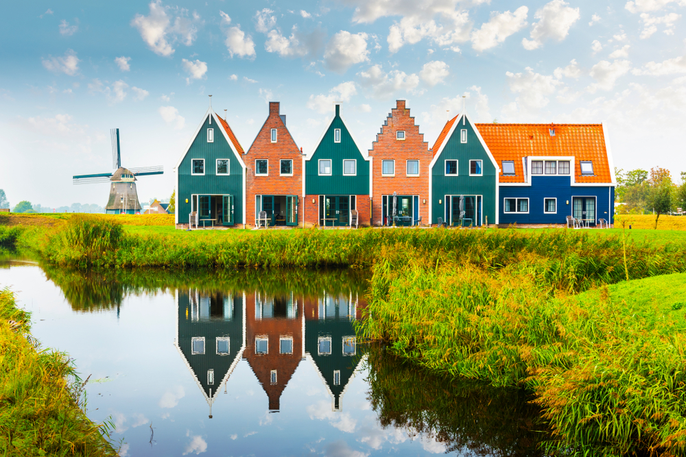 Simple vibrant houses by the side of a small lake with an old windmill at a distance, captured for a guide about the safety of visiting the Netherlands.