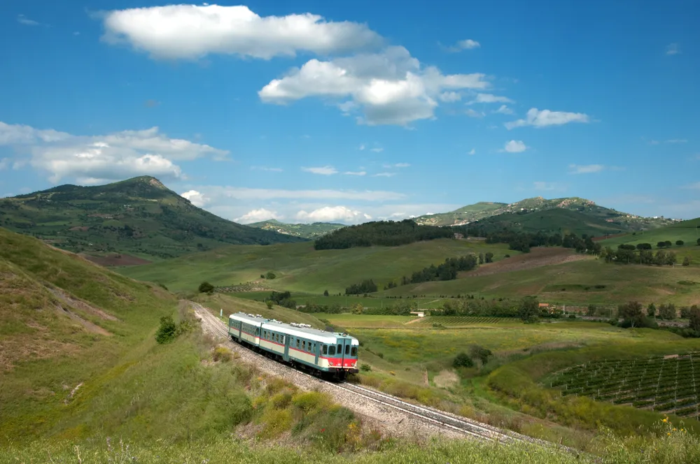 A short train travelling on a countryside with green fields and tall mountains, a section image for a travel guide about trip to Sicily cost.