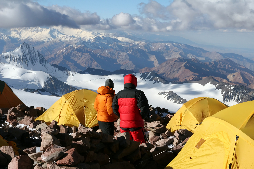 Two people camping on top of a rocky mountain, snapped as a section image for an article about trip cost to Patagonia, the surroundings are covered with snow due to high elevation. 