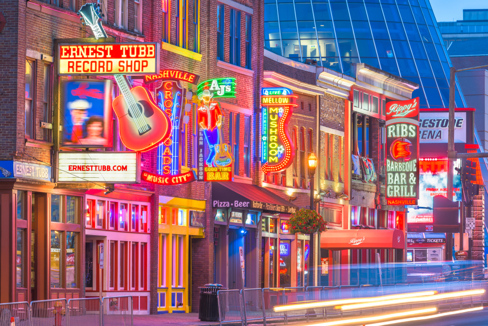 Storefronts of musical stores and bars at night illuminated by neon lights, captured for a piece on an article about trip cost to Nashville.