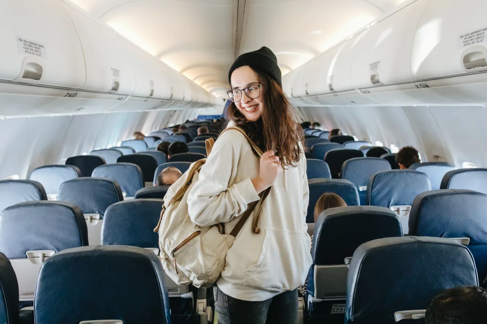 Young woman smiling in a hat and glasses wears a backpack as she boards the plane for a frequently asked questions section answering is a backpack a personal item?
