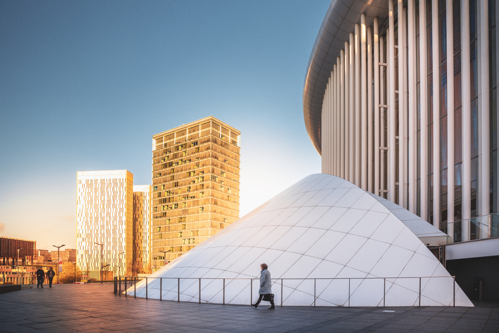 A person walking beside a structure with a beautiful architecture in Kirchberg, our pick on the best areas to stay in Luxembourg, other modern buildings is seen from a distance during a sunset.