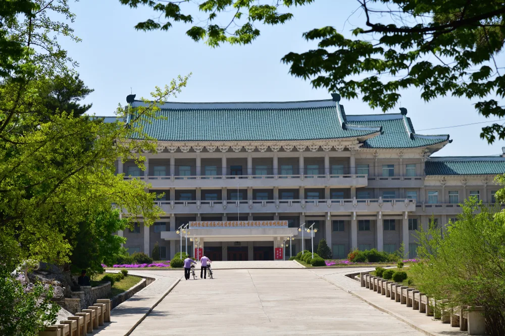 A large old building where two men can be seen walking towards it while pushing their bicycles.