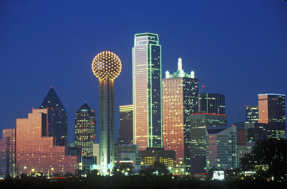 A city skyline at night pictured as a piece on an article about trip cost to Texas, one building has green lights on the edges, and one has a sphere on the top with lights.