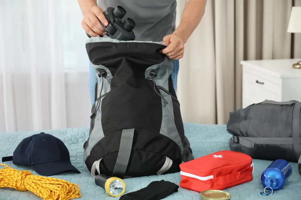 A man packing for a trip to Costa Rica, filling a backpack with binoculars, rope, hat, flashlight, first-aid kit, and tumbler.