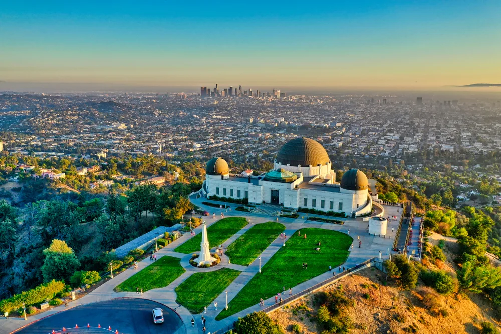 A structure with dome roofs built on top of a hill with a view of a large city during sunset.