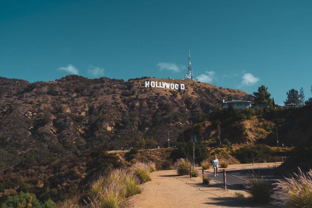 A woman walking on a trail with the view of the Hollywood sign on top of the hill. 