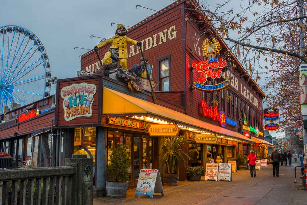 A building with several restaurants on its ground floor, for a piece on an travel guide about trip cost to Seattle, people are passing by beside the establishment and Ferris wheel is partially seen in background.