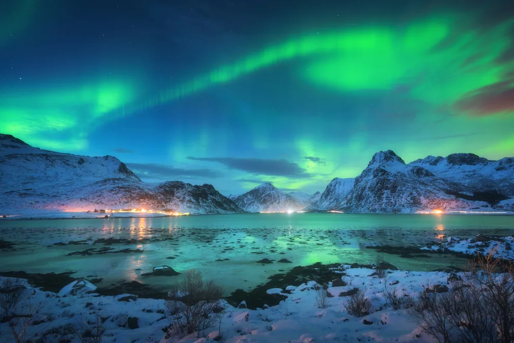 Beautiful northern lights where waves of green light can be seen in the sky and the ground and mountains are covered with snow, an image for a travel guide about trip cost to Scandinavia.
