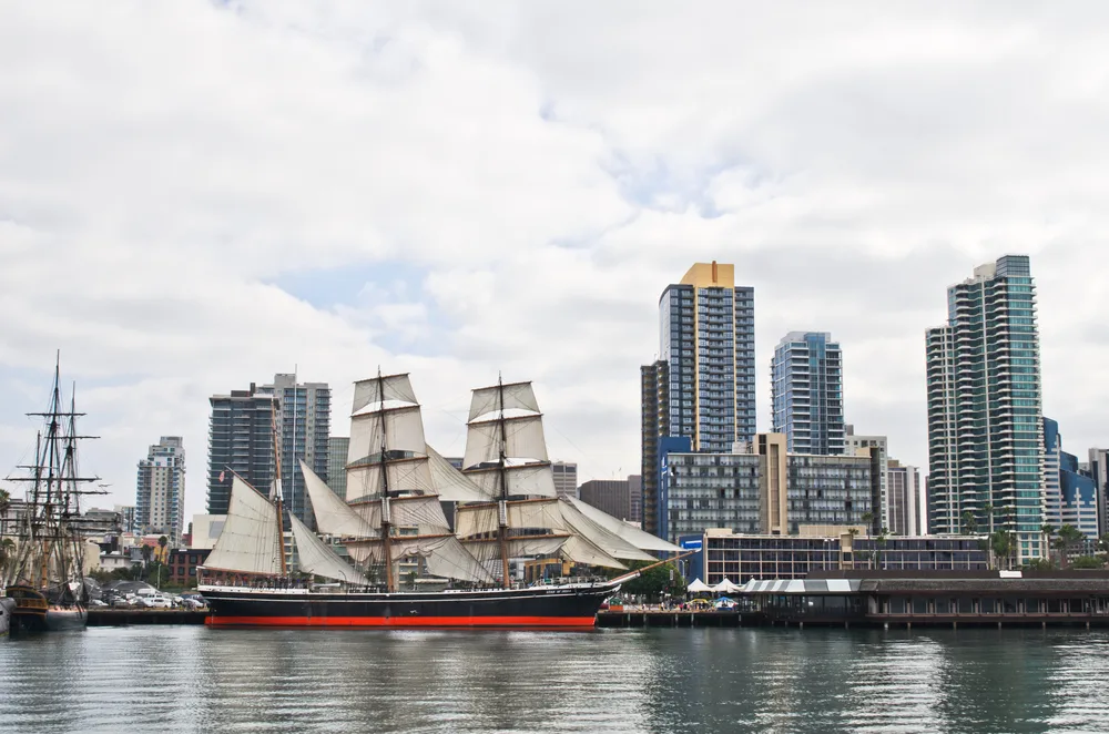 A large old boat with many sails on a pier near a city with tall buildings. 