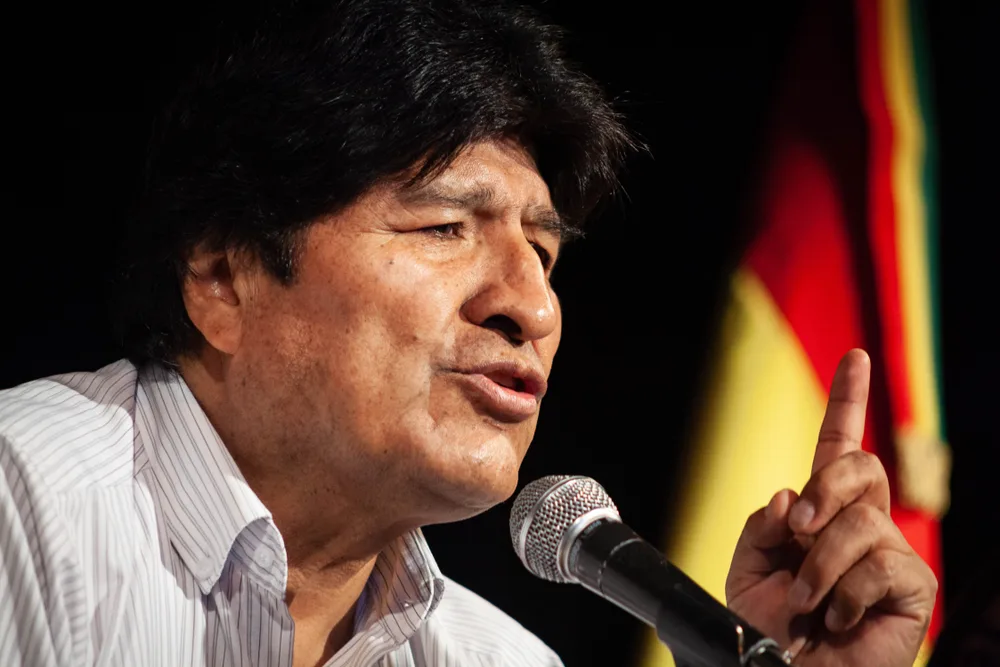 A man speaking on a microphone with his index finger pointing above, and a Bolivian flag in background. 