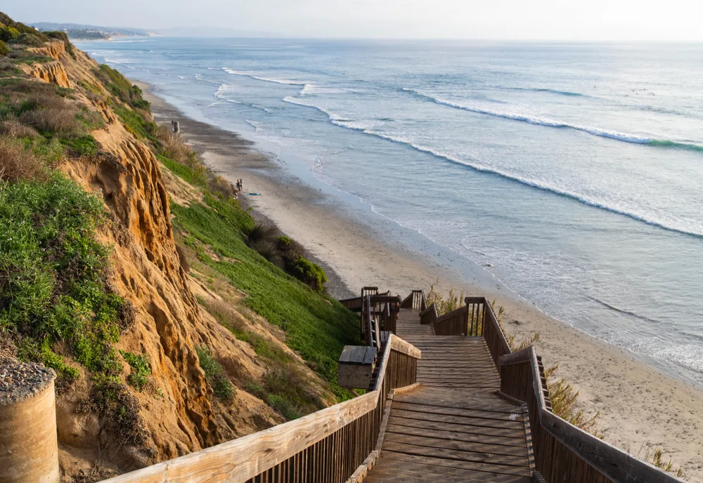 A wooden staircase leading down from the steep rocks to the beach during a calm afternoon, a piece on an article about trip cost to San Diego.