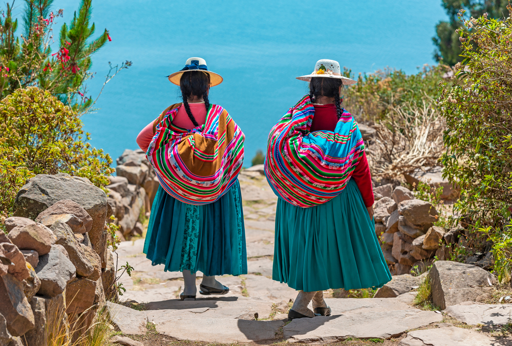 Two women wearing traditional clothes and hats, carrying their items inside a blanket wrapped on their shoulders while walking down a path. 