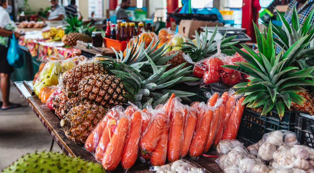 Fresh tropical fruits and vegetables displayed on a stall at a public market in Mahé, one of the best areas to stay in Seychelles, others stalls that sells bottled merchandise and costumers buying  is blurred in background.