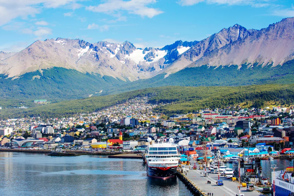 A populated town located between mountains and the sea, where a large boat can be seen docked on a pier. 