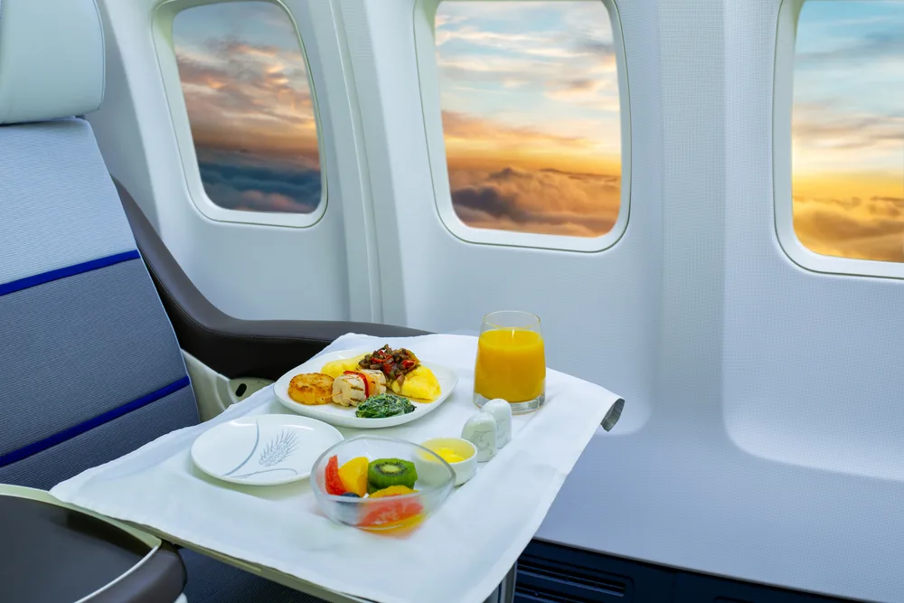Close-up of first class plane seat with flip-out armrest tray and breakfast food in the bulkhead front row with a sunrise outside the plane window