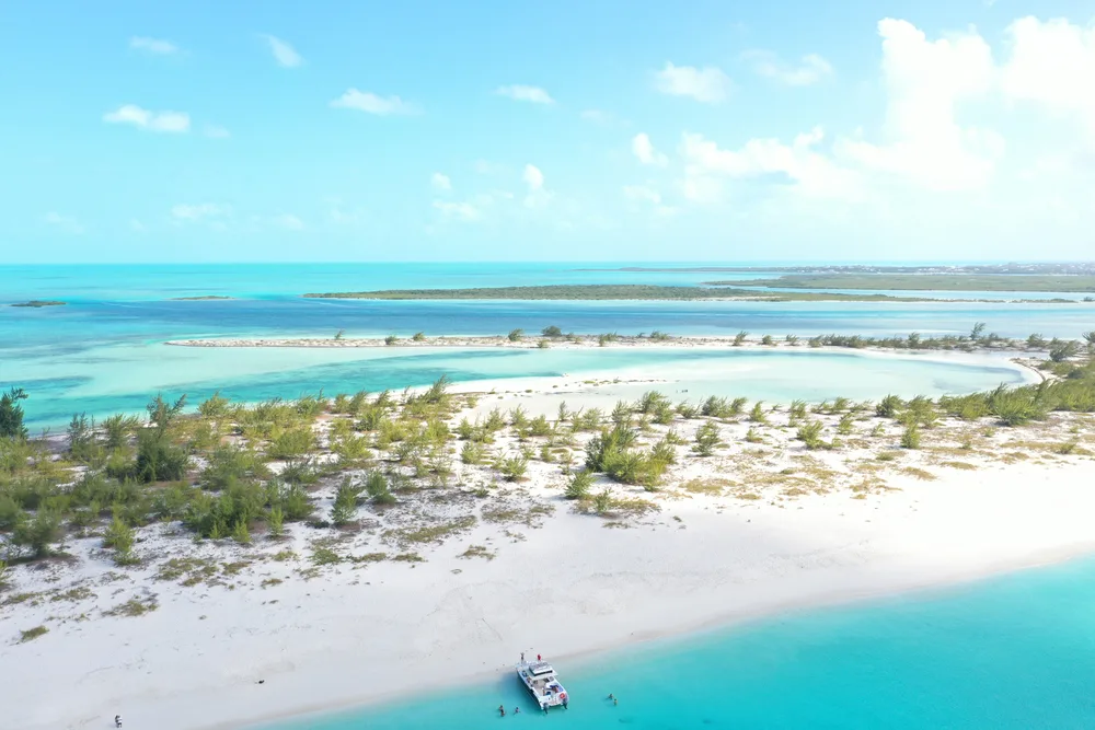 Stunning daytime view of a Turks and Caicos beach with scrub and white sand as a boat docks on the shore for a guide to average flight times to Turks and Caicos from the US