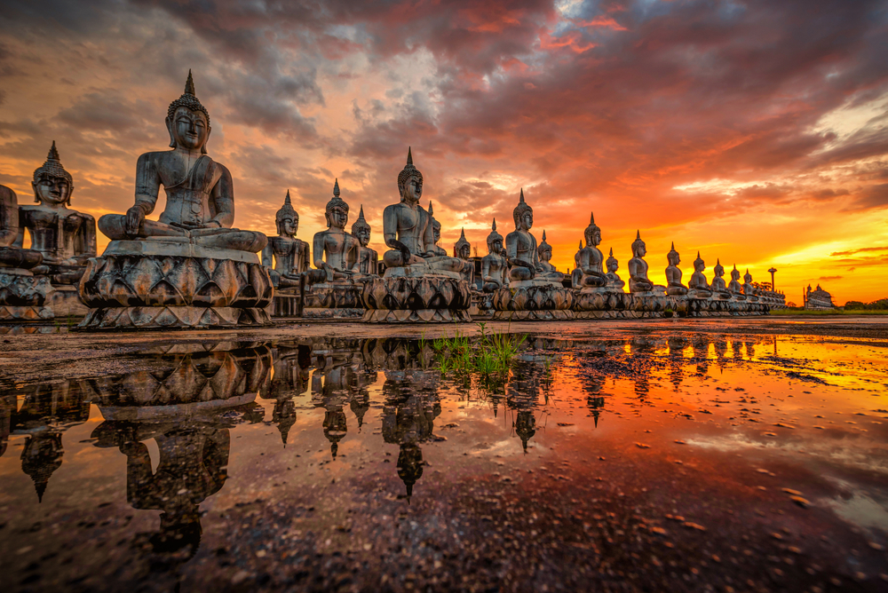 Row of aligned Buddha statues in Southern Thailand at sunset reflecting into the water shows what you'll see after the time it takes to fly to Thailand