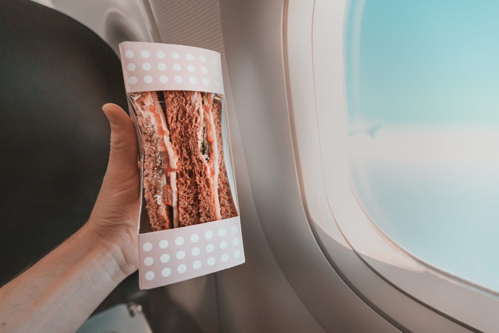 Man holds up a packaged sandwich in front of an airplane window to show the snack he brought on a plane