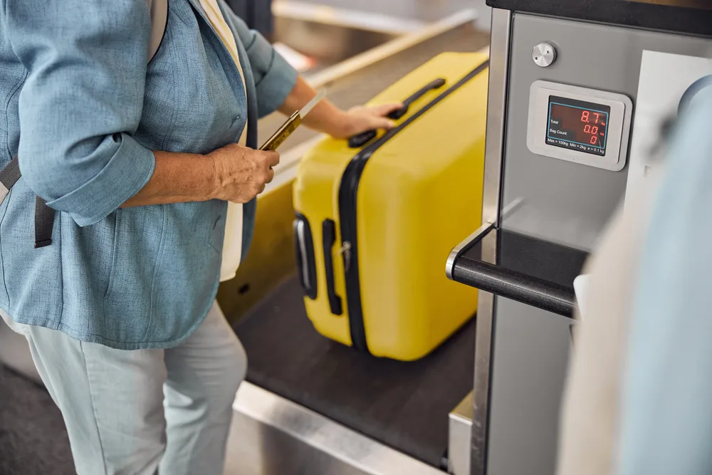 Woman places her yellow luggage on a conveyor belt at the airport to weigh and screen it before boarding to show the concept of Spirit Airlines fees for different types of baggage
