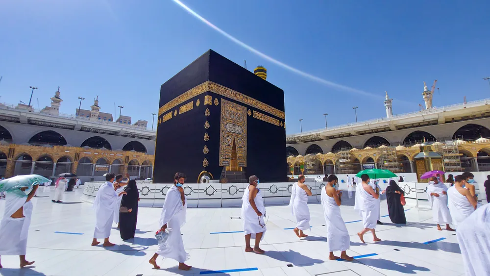 People walking barefoot in circles around a huge black box while wearing a traditional white Muslim clothing during a hot afternoon, an image for a travel guide about trip cost to Mecca.