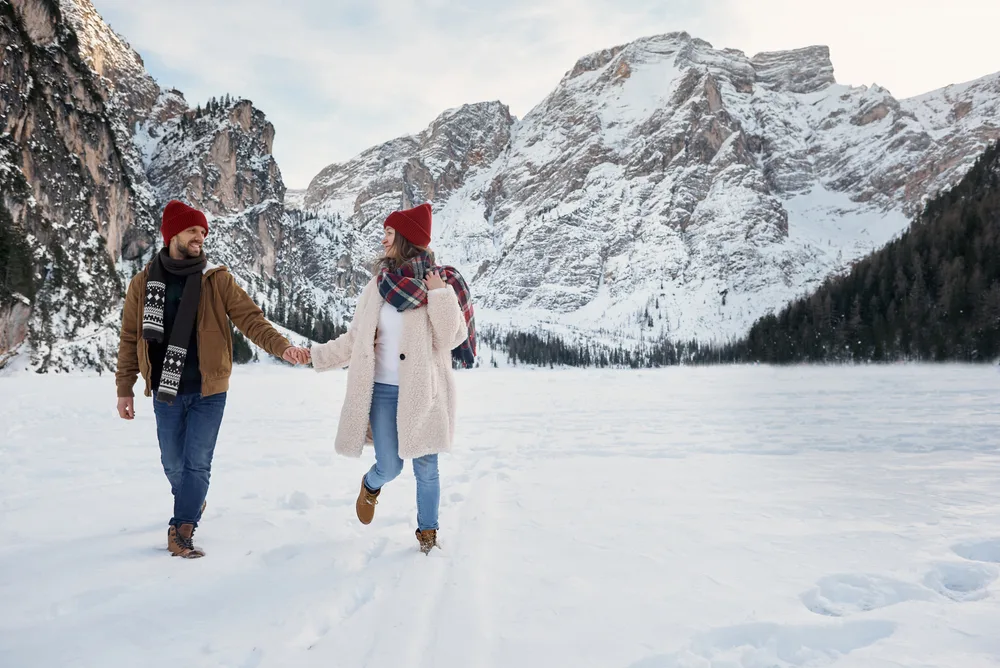 A couple walking on snow while wearing layers of clothes and icy mountains can be seen in background. 