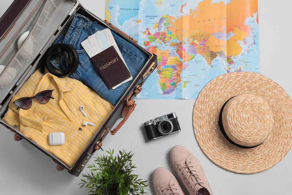 A suitcase filled with clothes with passport, sunglasses, earphones and a camera, shoes, hat, and a map outside the suitcase. 