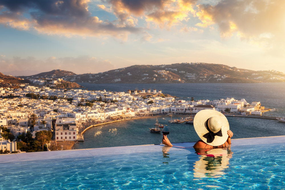 A woman swimming on an infinity pool with while holding a glass of wine and a summer hat overlooking a coastal town during sunset. 