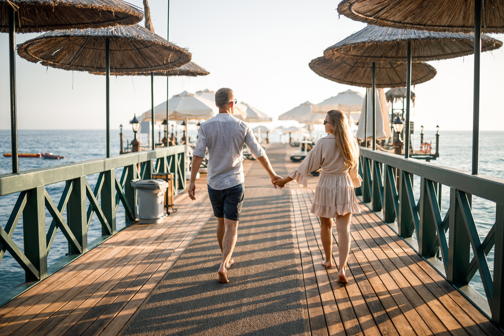 A couple walking while holding hands on a boardwalk with native umbrellas and the sea is visible in background. 