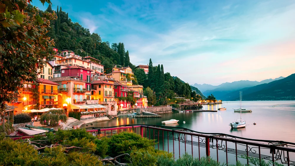 Houses built in front of a calm lake where a few small fishing boats can be seen moored on Lake Como for a guide to the best places to stay in the area