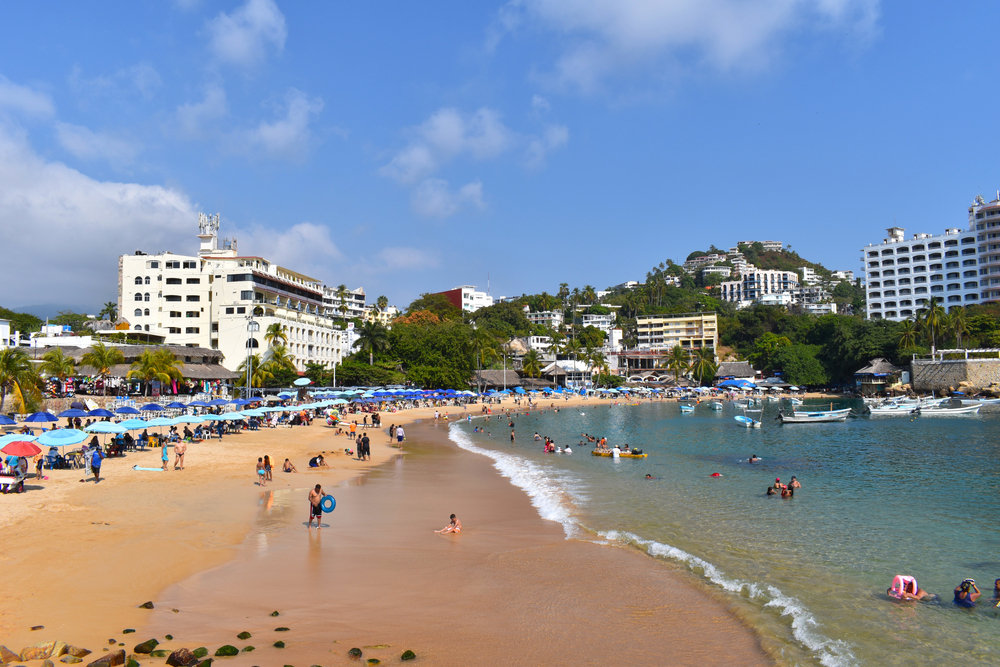 Many people swimming in the ocean by the dark brown sands during the best time to visit Acapulco on Caleta Beach