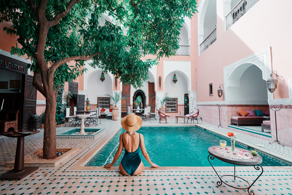 A woman wearing her swimwear, seating by the side of the pool in a hotel with a glass of drink placed on the table beside her, a concept image for a travel article about trip cost to Morocco.