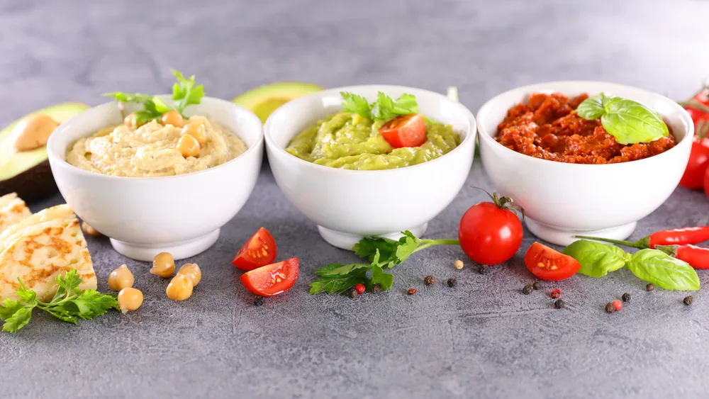 3 containers of sauces and dips with hummus, guacamole, and tomato tapenade arranged in white bowls to show what snacks are prohibited over 3.4 ounces on planes